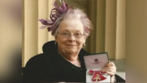 Oasis Norfolk Barbara Ross is wearing a black outfit and purple fascinator in her hair. She is looking directly at the camera and smiling and holding her OBE honour. 