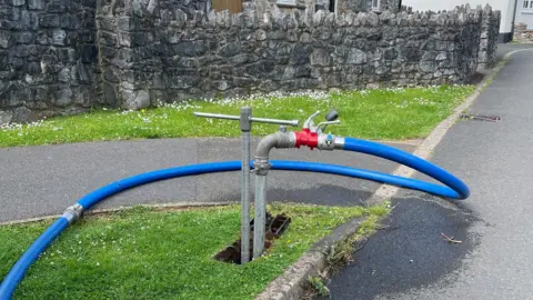 A standpipe with a tap and wide hose pipe leading from it across a footpath. Behind it is a wall and behind that is a stone house which has a conservatory.