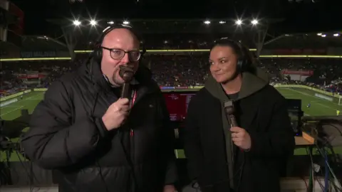 BBC Wales Sport BBC Wales football commentator Simon Davies, holding a microphone in the commentary box, alongside Nia Jones, also holding a microphone - both wearing coats - at half time during the match.
