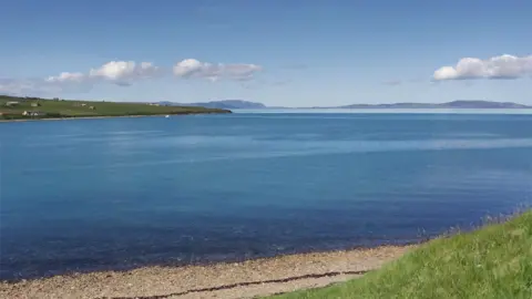 A large expanse of water. Some small hills in the background and a small pebble beach in the foreground. A blue sky with small, light clouds