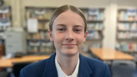 Freya has brown hair tied back and is wearing a blue blazer, white shirt and blue and yellow tie. She is sitting in a classroom full of books.