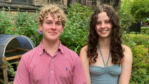 BBC/Seb Cheer A young man and young woman smile at the camera. They are standing in a garden, with plants behind them.