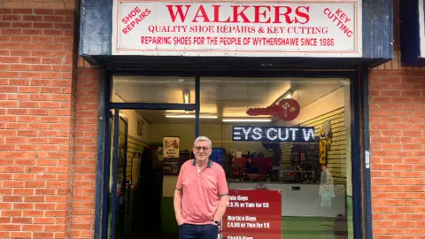 Gary Walker is standing in front of his shop, which has a large window. He has grey hair and glasses and is wearing a red t-shirt and dark jeans. A white sign is above the window reading Walkers in red writing. An scrolling LED light sign is in the window which, caught mid-rotation, shows part of the phrase "keys cut", but with the "k" missing.