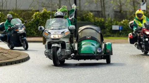 Roadrunnerpics A motorcycle with a sidecar going around a roundabout. The bike is silver, while the sidecar is painted bright green with a vintage look. Other motorcyclists are visible nearby. 