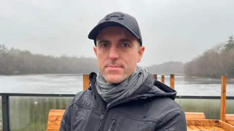 Mark is stood infront of the Bracebridge Pool in Sutton Park with some of the still-standing furniture in the background.