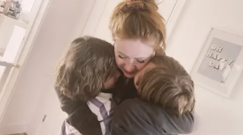 The Brothers Shed A woman with ginger hair hugging two young boys inside a house