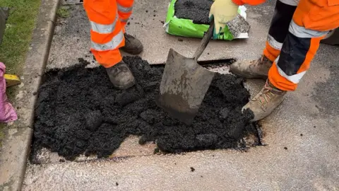 Chris McHugh/BBC Two workmen are seen filling a squared out area around a pothole with tarmac and levelling it out with a large shovel