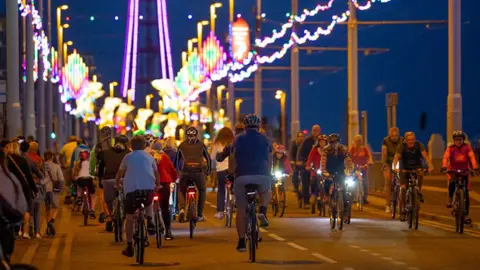 Visit Blackpool Blackpool prom at dusk with dozens of cyclists seen at a distance riding under Blackpool Illuminations