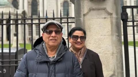 A man and a woman are standing outside railings, behind them is a grand looking white washed Belfast City Hall.
The man has a grey cap on, black sunglasses and he is wearing a black puffer jacket with a hood.
The woman has black hair, tied up, black square sunglasses on and a brown scarf on. She is wearing a black blazer.
They are both smiling at the camera.