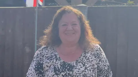 Jack Maclean/BBC Jane High a woman standing outdoors in front of a wooden fence. She has wavy brunette hair and is looking directly at the camera and smiling. She is wearing a black and cream coloured T-shirt and has a chain necklace round her neck.