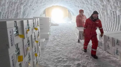 two people in sub zero protective red jump suits carry a large white chest between them through an icy tunnel towards the camera. Lots more similar boxes are stacked on each side of the walls of the cave.