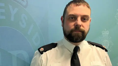 Leanne Harper/BBC Supt Tony Fairhurst, who has short brown hair and a brown beard and wears a police uniform of a white shirt, black tie and black shoulder epaulettes, looks into the camera against a wall bearing the crest of Merseyside Police.