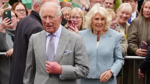 King Charles III and Queen Camilla during a public appearance. The royal couple are walking outdoors, surrounded by a crowd of onlookers who are taking photos and videos with their phones. King Charles is dressed in a light grey plaid suit with a patterned tie and pocket square, while Queen Camilla wears a pale blue outfit adorned with a small brooch. 