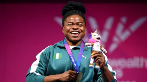Esther Nworgu smiles broadly during the medal ceremony for the lightweight para-powerlifting at the 2026 Commonwealth Games. Visible from the waist up, she wears a green and white Nigerian tracksuit and uses her right hand to clutch her gold medal, which is on a purple and pink ribbon around her neck, and her left to hold up a cuddly toy Games mascot. Blurred in the background is a red board with a white Glasgow 2026 logo on it