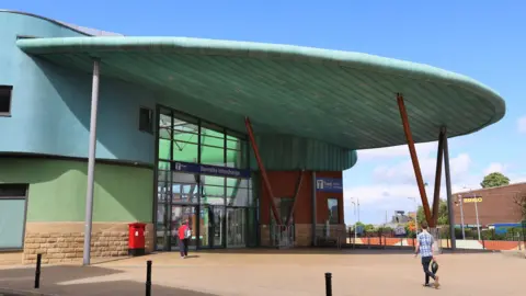 Barnsley Interchange. An external view of the interchange, a large semi-circular canopy supported by a number of struts extends from the double-height glass frontage of the building. There are a couple of people walking towards the doors.