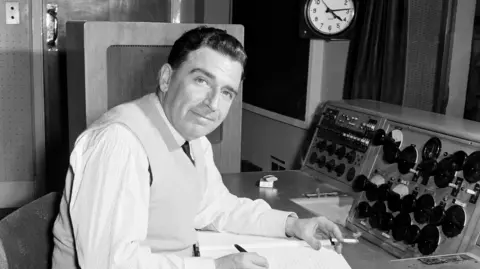 A black and white picture of Tony Shryane, who is looking at the camera sat in a radio studio with a pen in his right hand and a book open. He has a lit cigarette in his left hand 