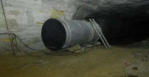 HSE Large industrial ventilation fan mounted inside a wide underground tunnel or mine, with a metal ladder leaning against the wall beside it and cables running along the ground in a dimly lit environment.