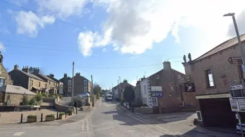 The main road leading through a sunny village. Stone properties are situated on both sides and there are roadsigns too.