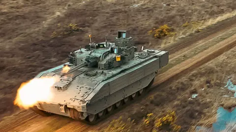 General Dynamics An Ajax armoured vehicle being driven through a muddy terrain. It's firing its gun.