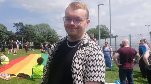 Curtis Lee has short blonde hair with dark bits on the side. He is wearing clear rounded glasses, a white and black shirt, a black t-shirt and a silver necklace. In the background is people standing and waiting for the pride parade to start, and there is a rainbow coloured flag.