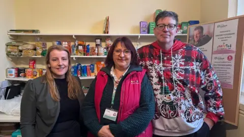 Andrew Turner/BBC Laura Rawnsley, on the left, has long blonde hair. She is wearing a black jersey and grey jacket. Next to her is Jemma Jackson, the operations manager of the Waveney Food Bank, wearing a blue knitted jersey and pink gilet. She has burgundy-coloured hair. Matthew Scade, the food bank project manager, is wearing a brightly patterned Christmas-them jersey. He has medium length dark hair and wears glasses.
