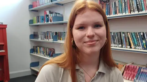 Debbie Tubby/BBC A teenage girl with long red hair down her back. She is wearing a beige polo shirt and is starting to smile. She is sitting in a library and behind her are shelves with books. 