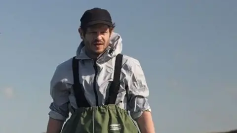 Iwan Rheon standing thigh deep in water wearing green waders