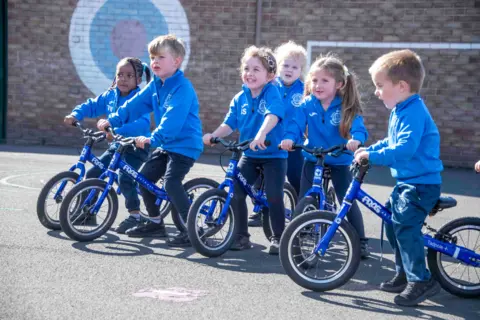 Six primary school pupils in bright blue jumpers sit on blue bicycles in school playground