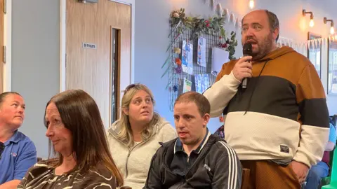 A man with a beard speaking into a microphone held close to his mouth. He is standing up with people sat next to him turning their heads towards him and listening.
