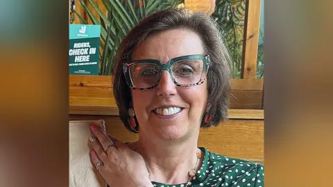 Handout Emma Martin with brown-bobbed hair wearing dark green and leopard print glasses and a dark green and white polka-dot top sits in a cafe. She is smiling. 