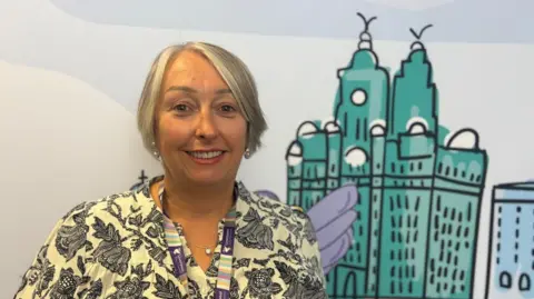 BBC/Claire Hamilton A woman with short grey hair smiles as she looks directly at the camera. She is pictured in front of a brightly coloured cartoon of the Liver Building. 