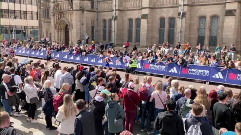 Hundreds of people line both sides of Oxford Road behind barriers as they watch runners pass through on the way to the finish. The old stone main building of the university with arched windows can be seen behind them.