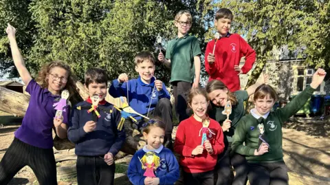 A group of children wearing different coloured school jumpers smile at the camera, all holding wooden spoons that have been decorated to look like their favourite book characters.