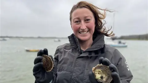 Lottie Johns who has brown hair standing on a pontoon with the harbour and yachts behind her holding two oysters 