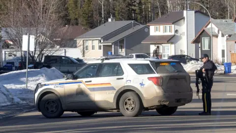 Policeman stands beside police car parked half across a residential road with mountain behind in Tumbler Ridge, British Columbia