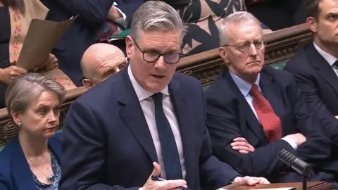 PA Media Keir Starmer speaking in the House of Commons. He has grey hair, wearing glasses, a navy blazer, white shirt and navy tie. Other MPs are behind him.