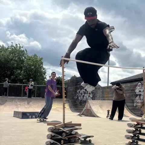 Angelle Joseph/BBC Joe Bourne, a skateboarder wearing all black who is jumping over a stick and landing on his skate board.