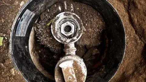 Yorkshire Water A hedgehog which is stuck in a water pipe. It is curled around a section of the pipe and there is soil around the outside.
