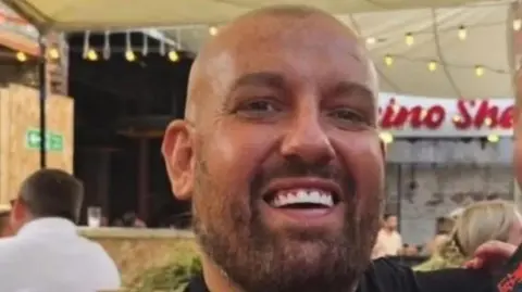 Close up shot of Stephen Cavanagh. His is bald, tanned and has bright white teeth. The backdrop appears to be a bar or food court.