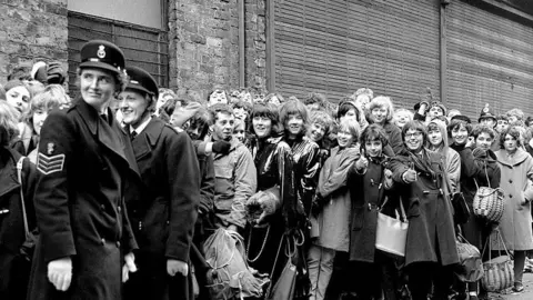 Getty Images Uniformed women help to keep crowds of teenagers back who are queueing for tickets for the Beatles at Liverpool Empire Theatre