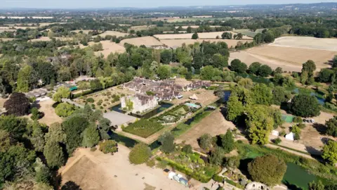 Hever Castle and its grounds, seen from above. Amongst the manicured, green gardens are patches of yellow grass.