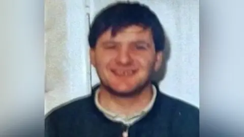 Family Handout An old photo of Paul Thompson. It is slightly out of focus. He is smiling at the camera. He has dark hair which falls over his forehead.  He is wearing a dark blue top with a white collared shirt underneath, 