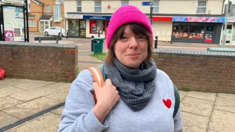 Sam Read/BBC Joanne Ollier, standing outside a row of shops that are behind her. She is wearing a bright pink hat, a blue and black scarf, a blue jumper, with a red heart to the right. She has one arm on her shoulder, holding on to a bag strap that is cream and orange.