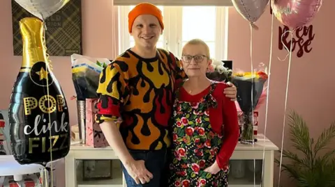 Contributor photo A man in an orange beanie and orange, red and yellow jumper standing next to an older woman with short hair.