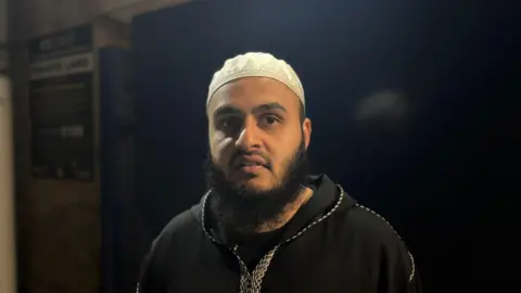Shariqua Ahmed/BBC Osama, a man with a beard, wearing a white skull cap and a black top.