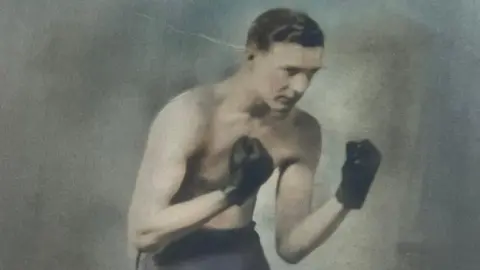 Family photograph An old photograph of Maynard Powell wearing boxing gloves. He has short dark hair and is not wearing a shirt. 