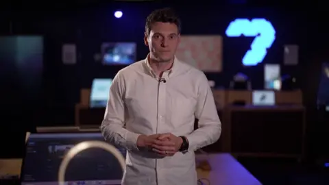 Joe Tidy standing in front of screens with his hands clasped in front