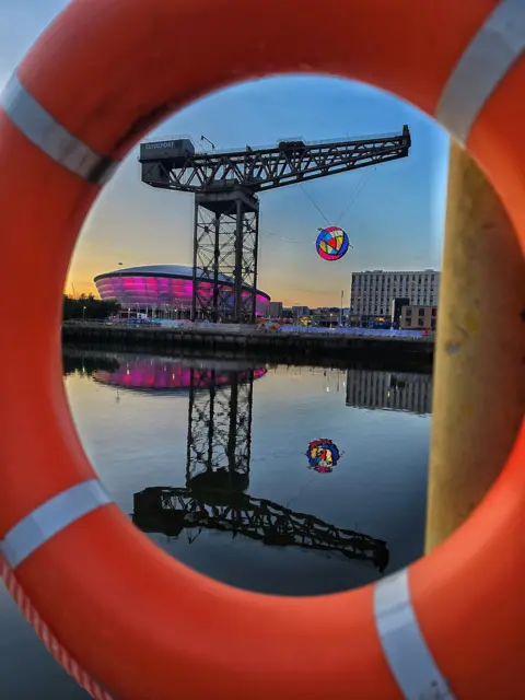 Eddie Romeo A large orange lifebuoy frames a view across the River Clyde. Through the centre of the ring, the Finnieston crane can be seen with the Commonwealth Games rose sculpture raised in the air and the Hydro illuminated in pink in the background.
