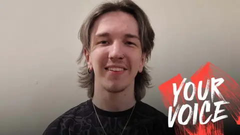 Liam Davidson Liam Davidson with blonde hair wearing a black t shirt with a grey pattern a silver necklace and a black nose ring and earring smiling at the camera with the graphic Your Voice