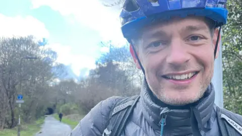 Euan Baxter is wearing a blue cycling helmet. He is smiling at the camera. The Roseburn Path is in the background.